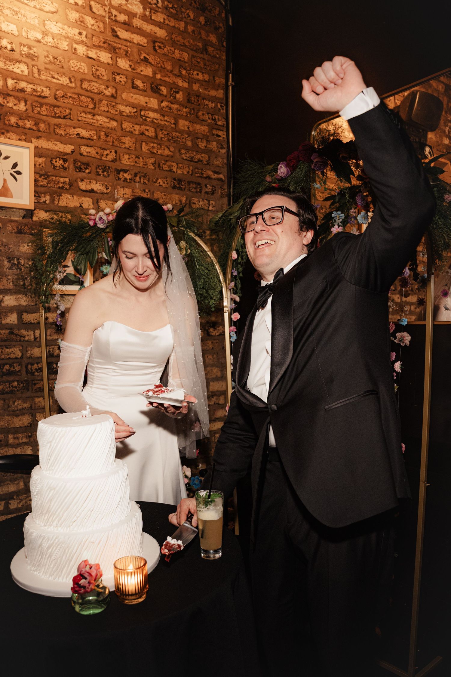Joyful groom raises fist in celebration while bride cuts white three-tier wedding cake at rustic brick wall venue.