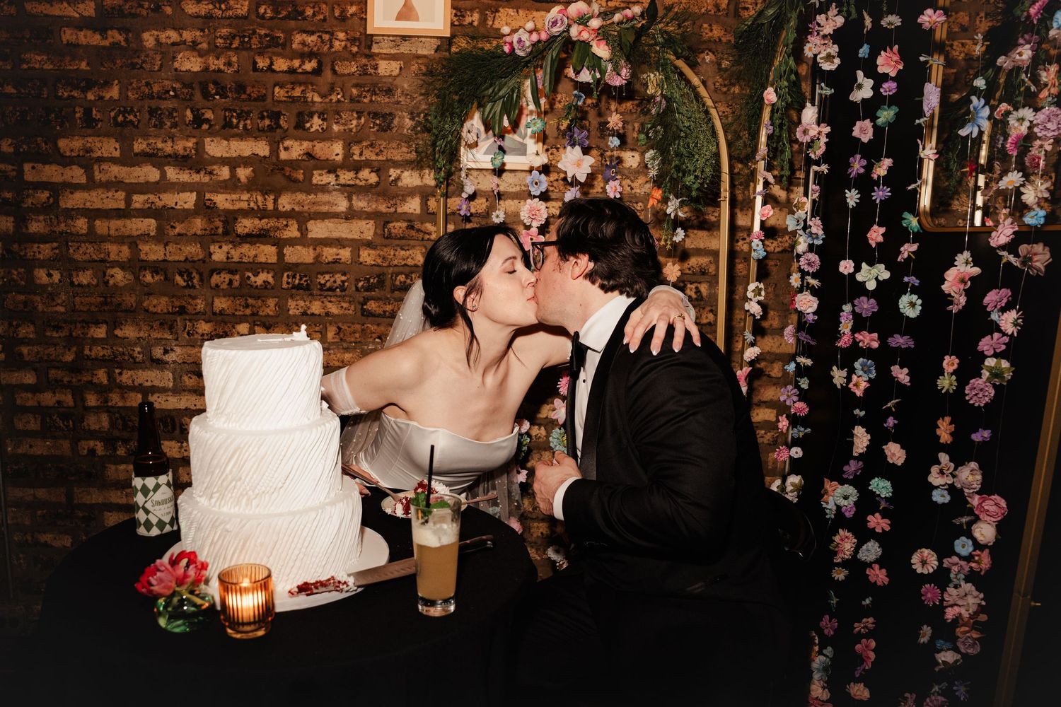 Newlywed couple shares romantic kiss behind elegant white three-tier wedding cake at reception with floral backdrop.