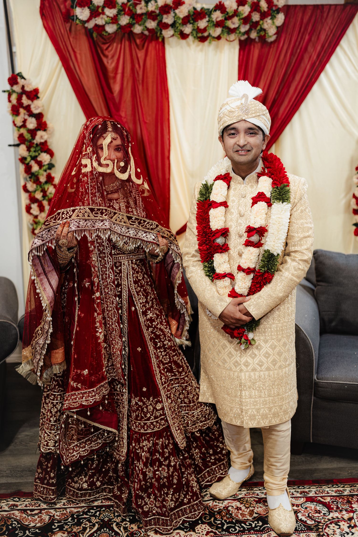 South Asian bride and groom in traditional red and cream wedding attire standing before floral backdrop at ceremony.