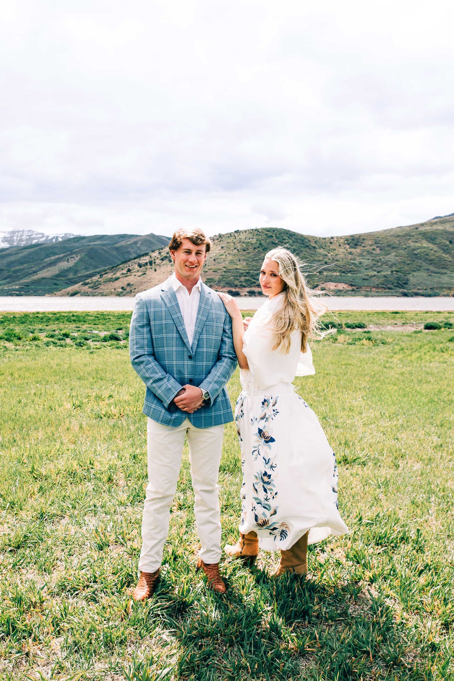 Couple posing outdoors in a green field with a lake and hills backdrop, man in plaid blazer, woman in floral dress.