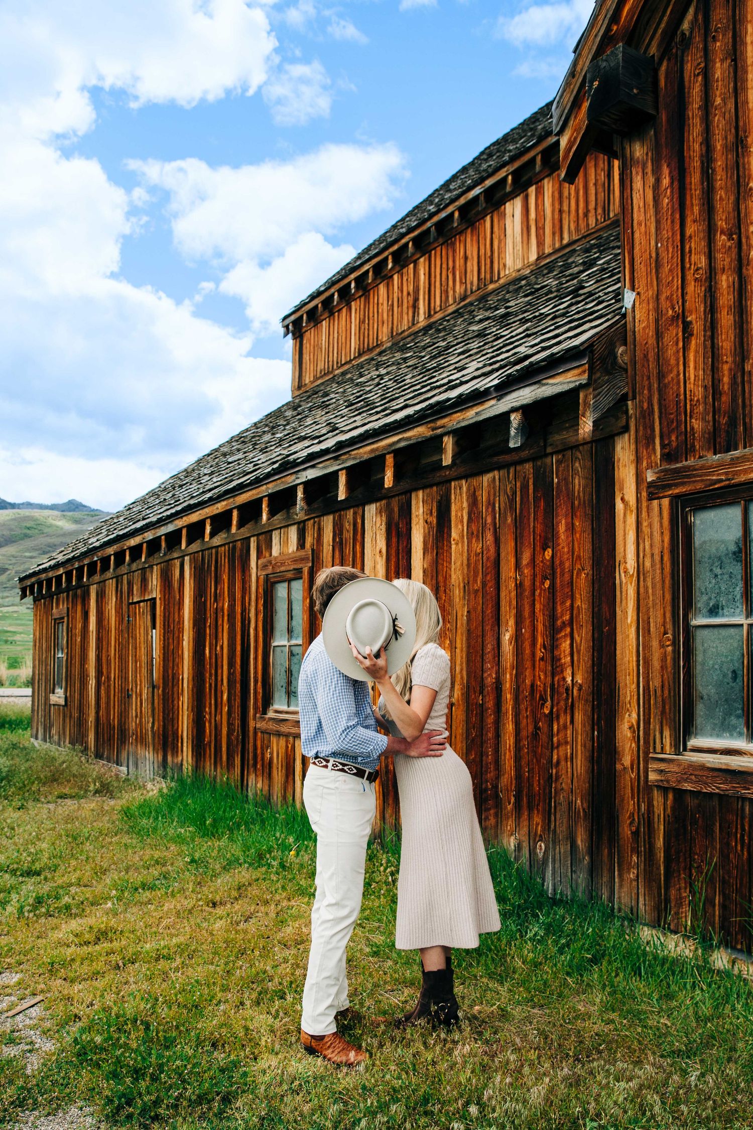 Couple kissing behind a white hat beside a rustic wooden barn under a blue sky.