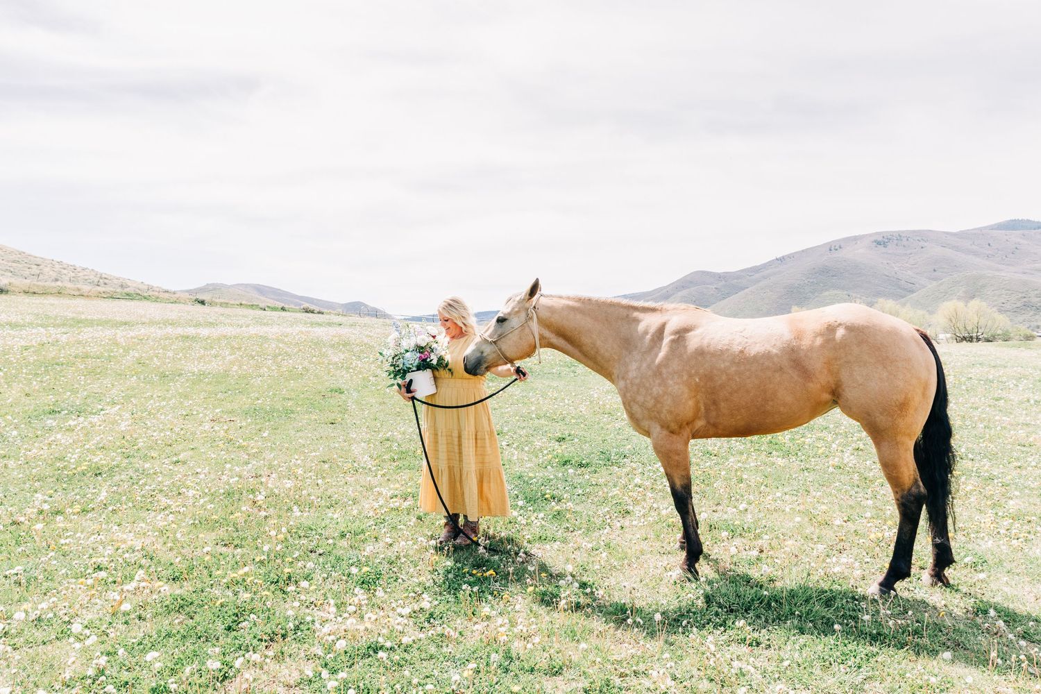 Bride in yellow dress holds bouquet while a tan horse nuzzles her in a green mountain meadow.
