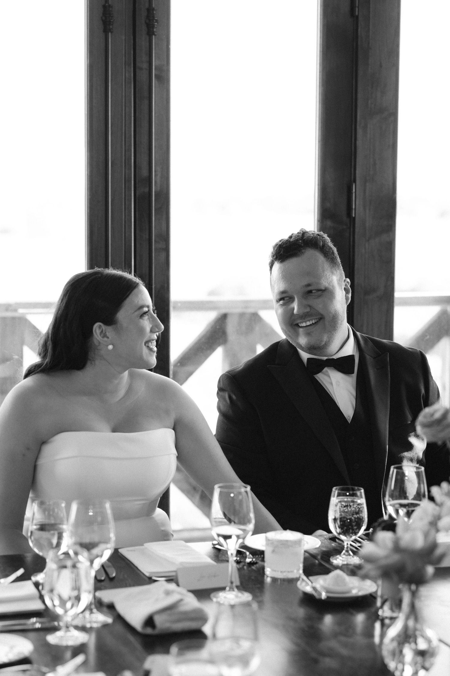 Black and white wedding photo showing a couple walking on grass with a brick house and evergreen trees in the background.