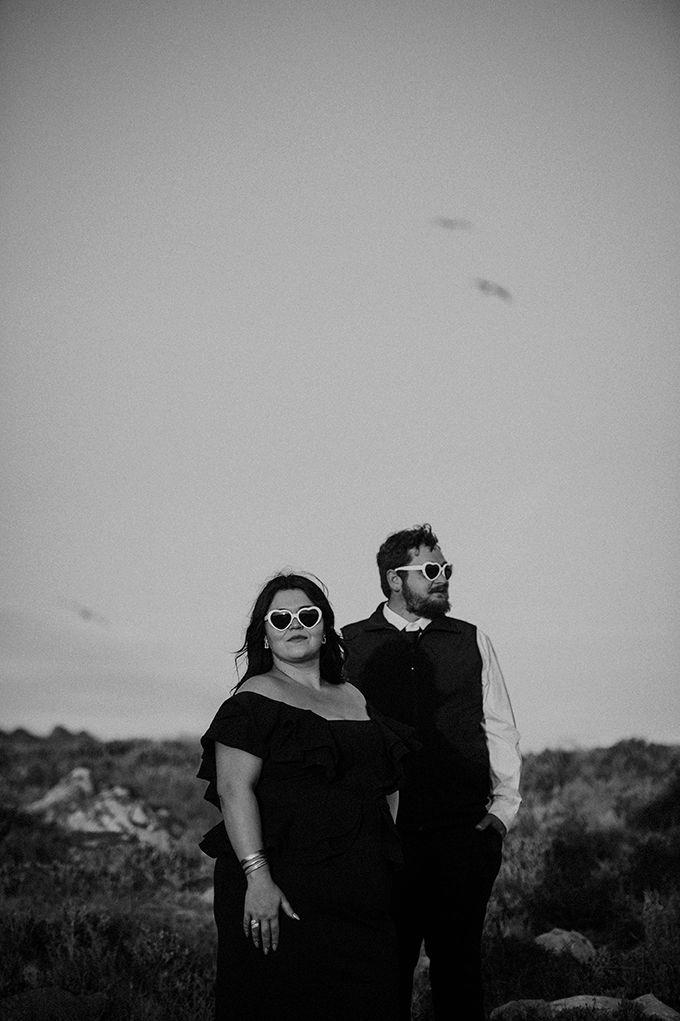 Elegant photograph of a couple dressed in black standing in each other’s arms against a rocky coastal backdrop at Fransmanshoek