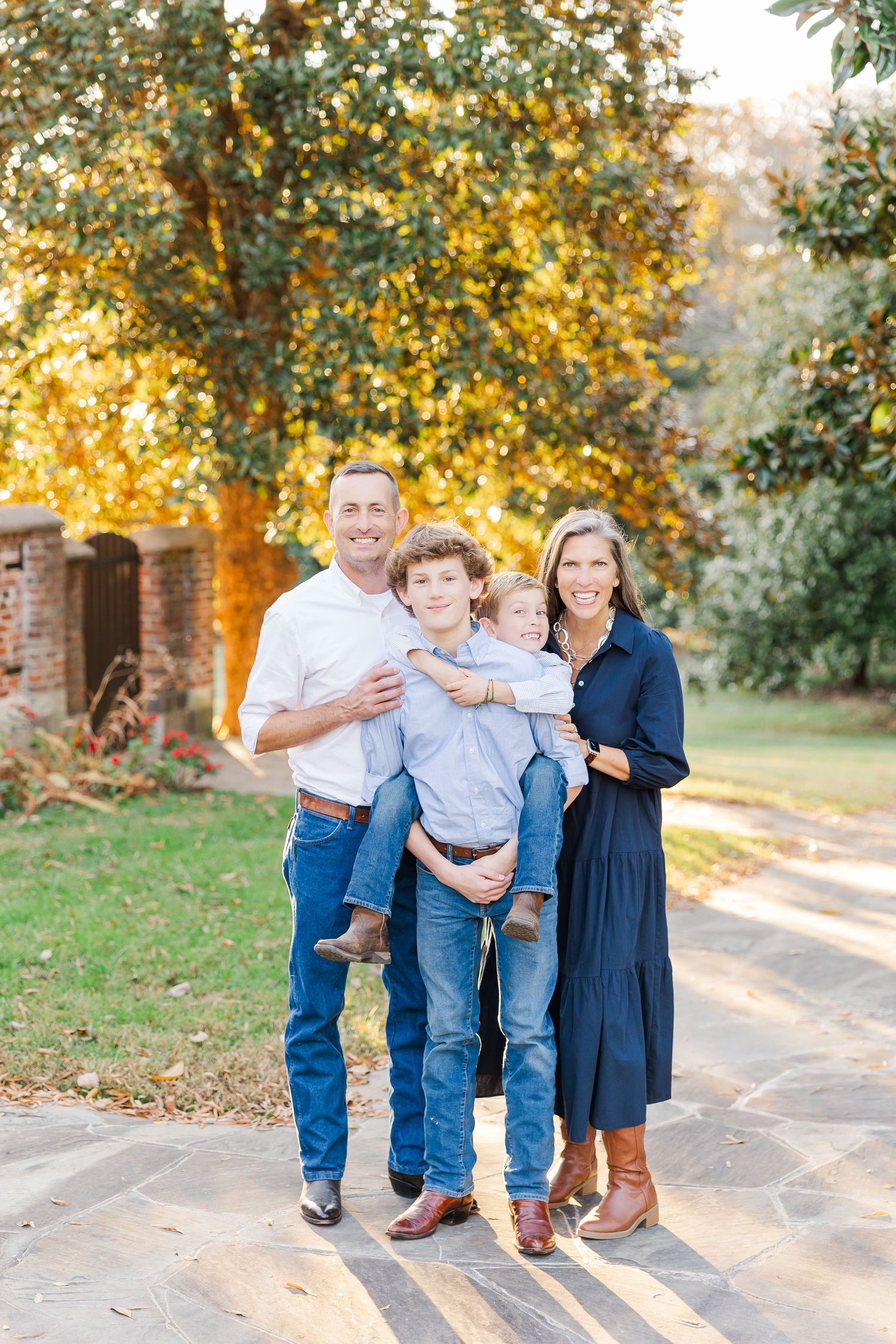 A family poses together outdoors on a path with autumn trees and golden sunlight in the background.
