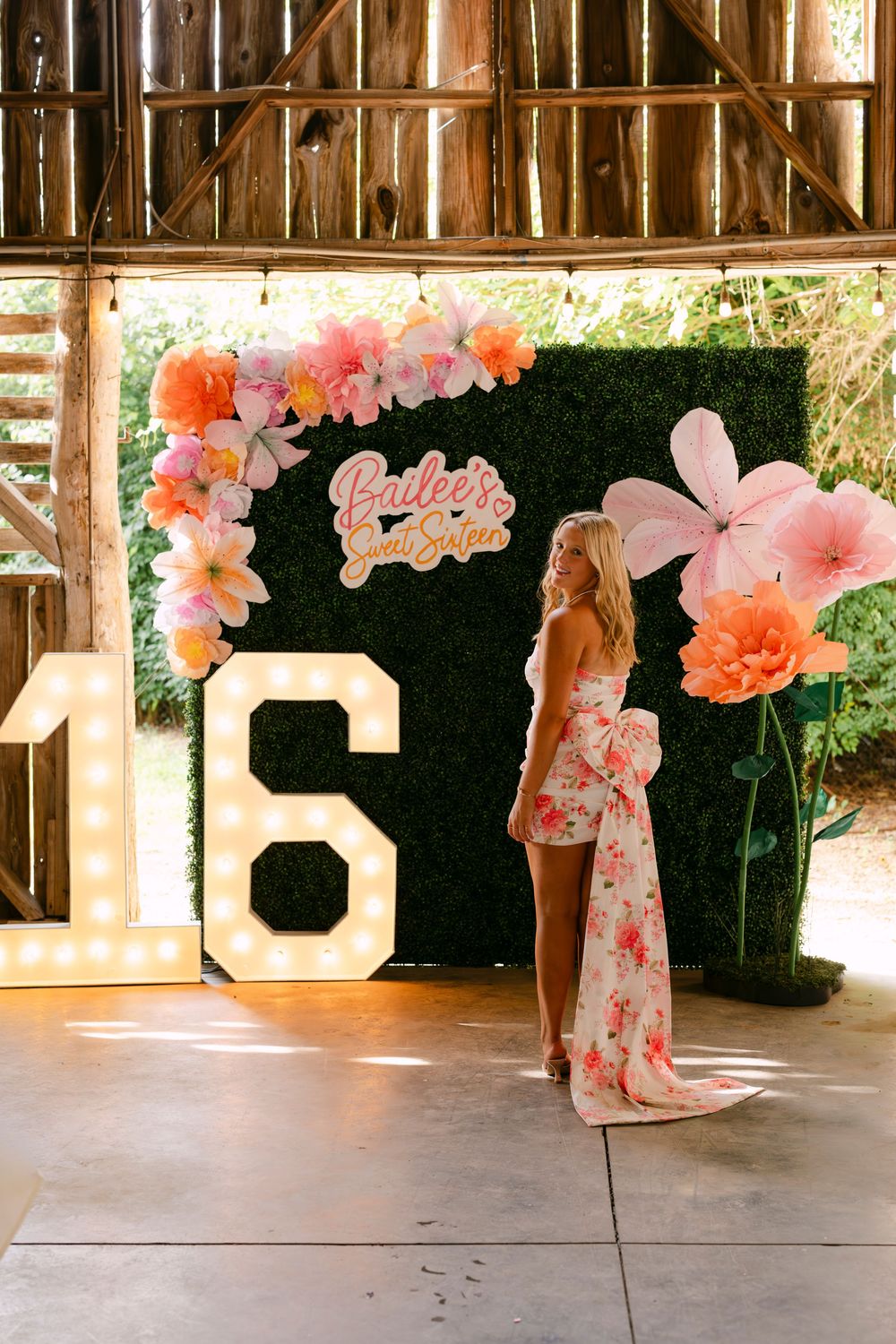 Sweet 16 birthday party setup with marquee numbers, floral backdrop, and a girl in a floral dress posing in a barn venue.