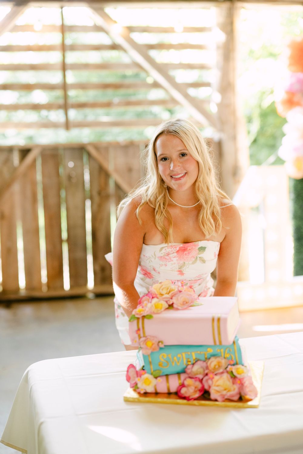 Young blonde woman in floral strapless dress smiling behind a decorated Sweet 16 birthday cake.