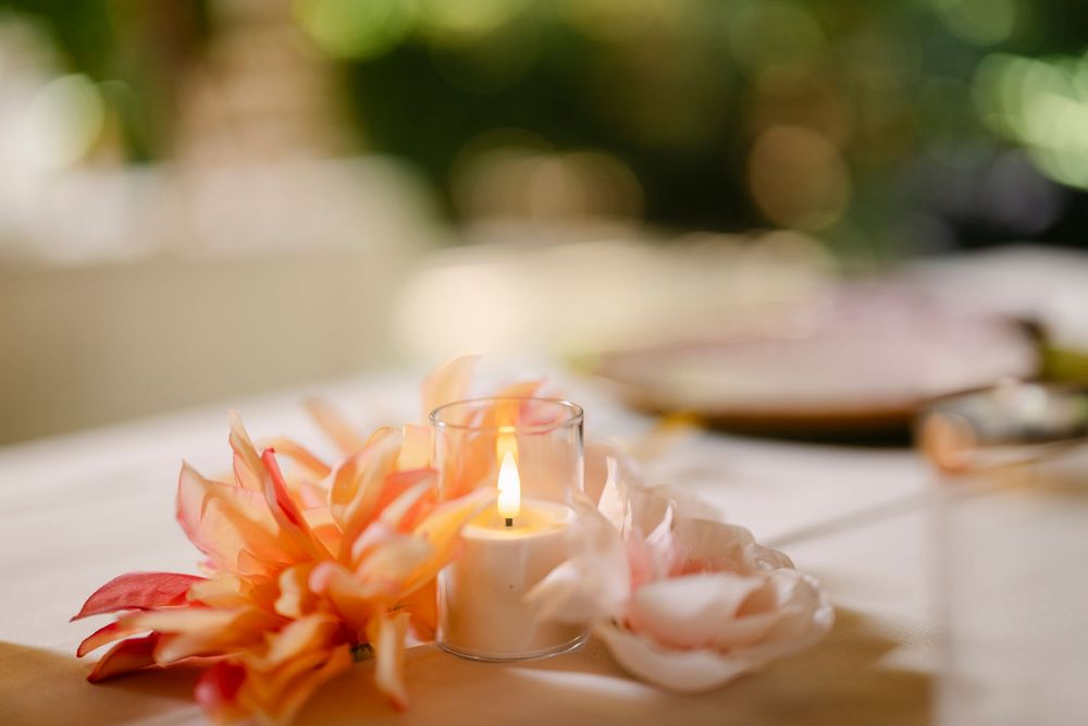 Lit candle in glass holder with pink tropical flowers on white table at outdoor wedding reception.