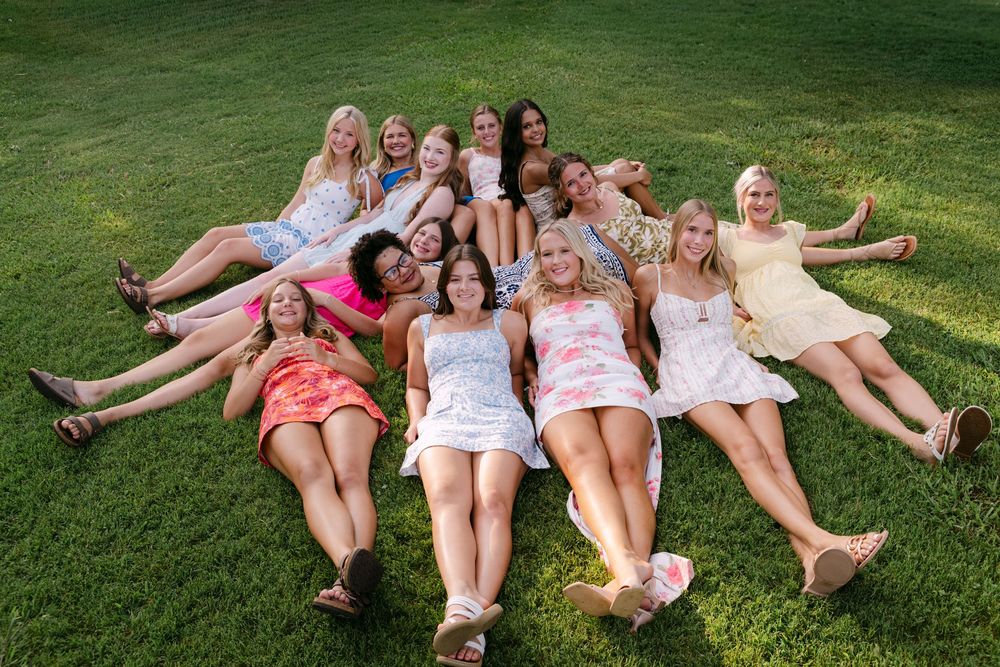 Group of teenage girls in summer dresses lying on green grass smiling and posing for a fun outdoor photo.