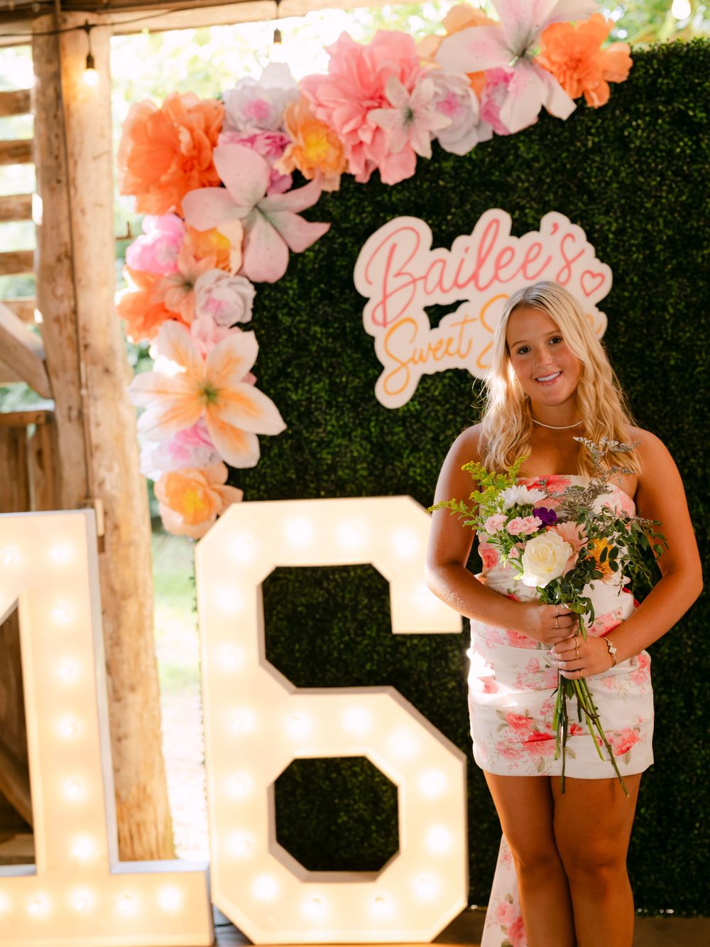 Teen girl holding flowers poses at Sweet 16 party with marquee numbers, floral arch, and hedge wall backdrop.