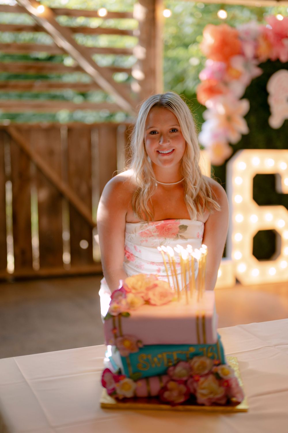 Smiling blonde woman in floral dress celebrating sweet 16 birthday with lit candle cake and marquee number decoration.