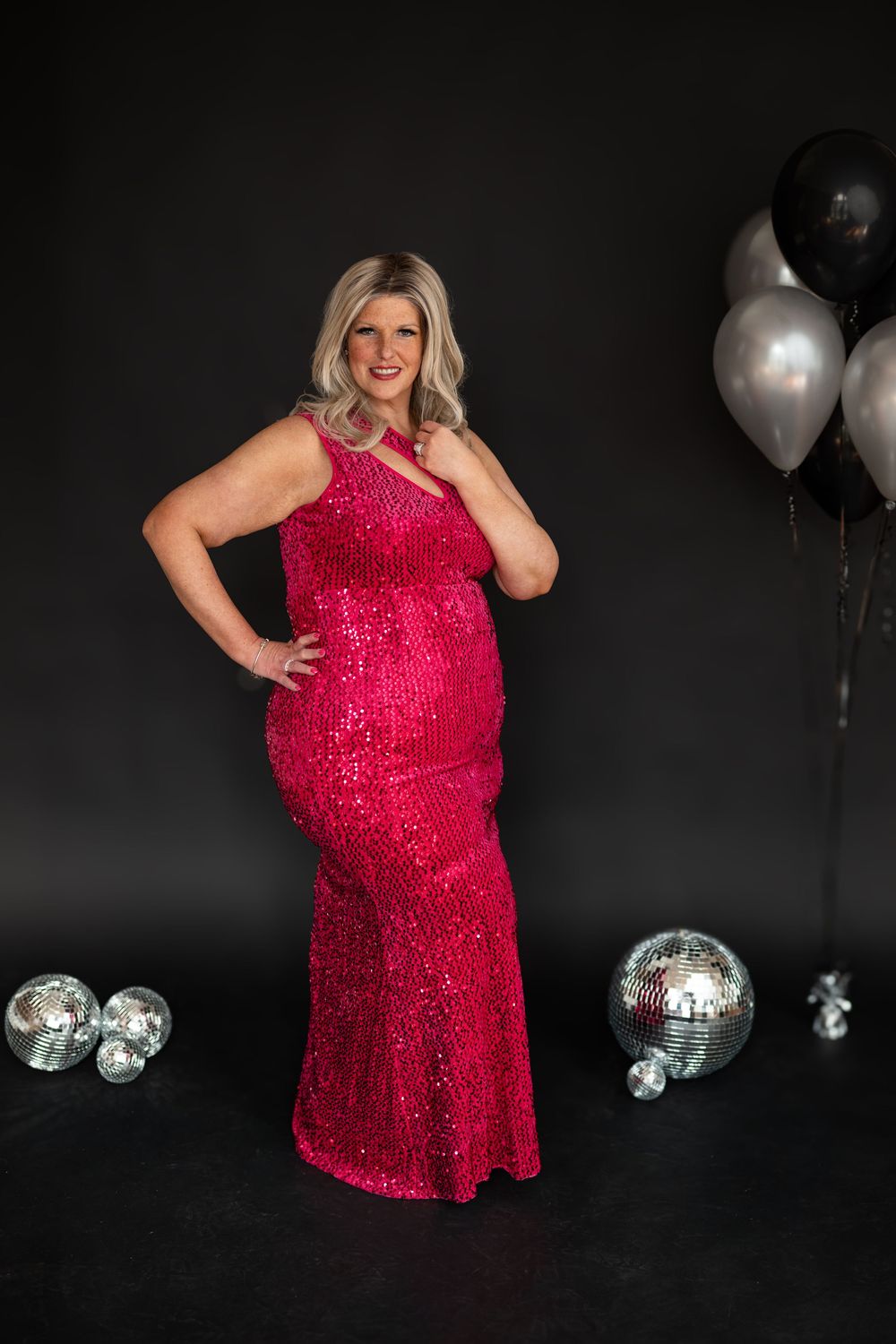 Plus-size woman in a pink sequin gown poses with disco balls and balloons on a dark background.