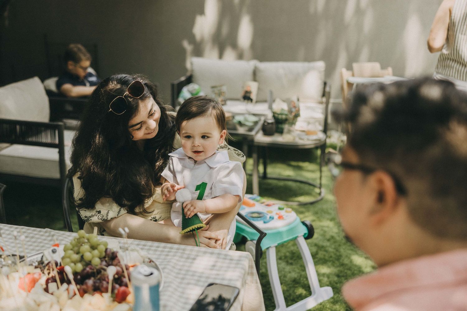 A family gathering outdoors at a table with drinks and decorations while holding a small baby in a highchair.