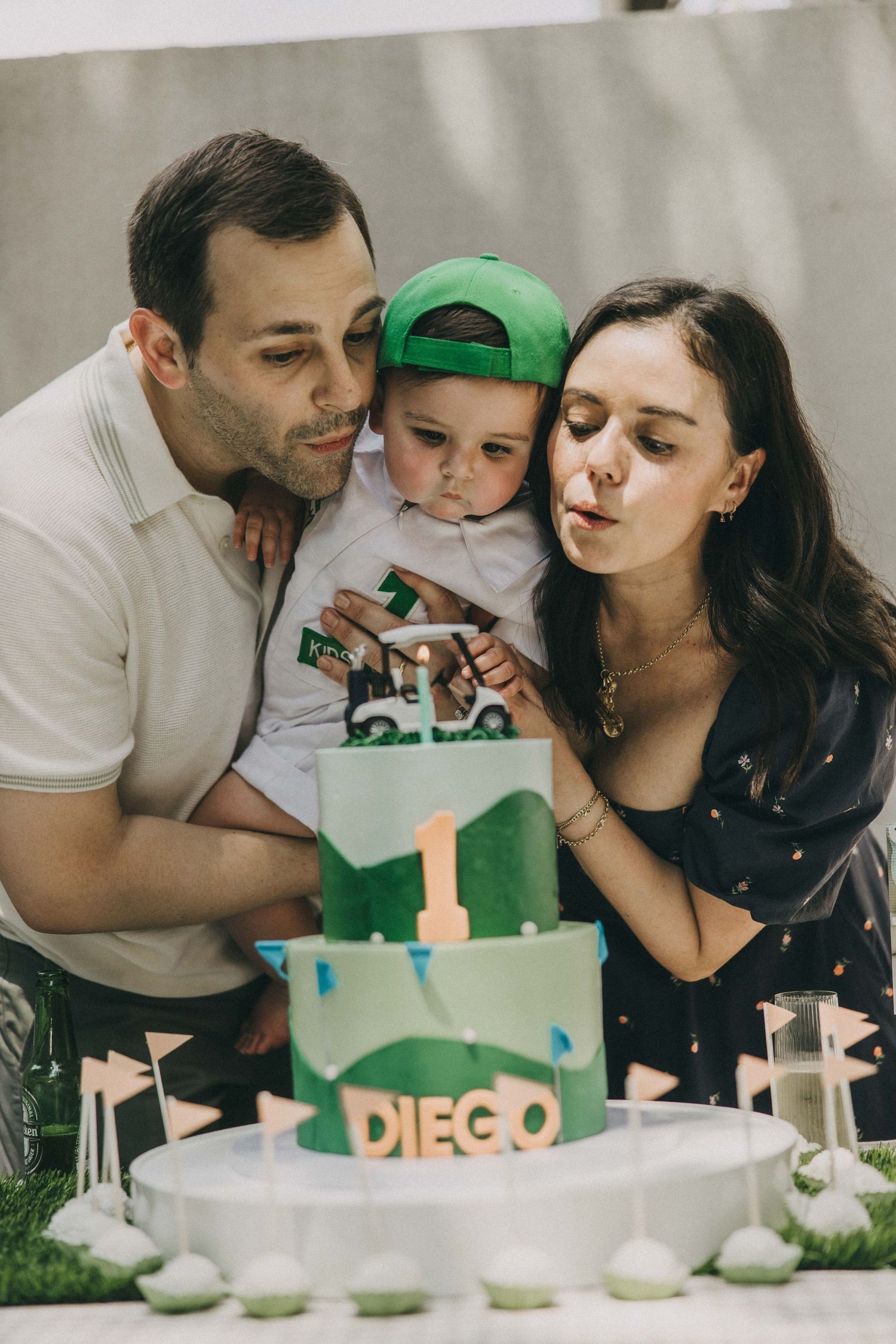 Family celebrates child's first birthday with green and white dinosaur themed cake at outdoor party.