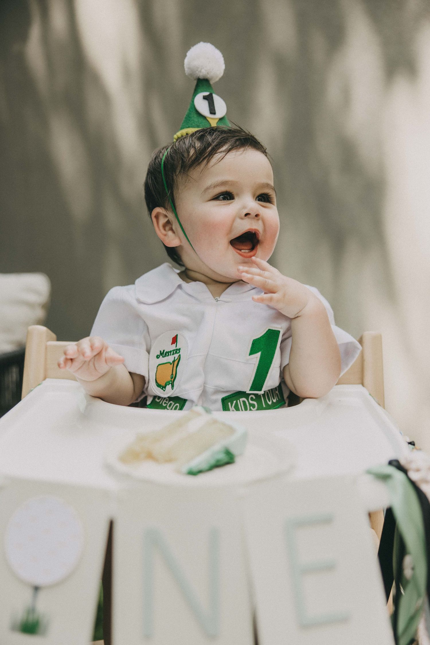 Baby wearing white bib with green accents in high chair making funny expressions while eating.