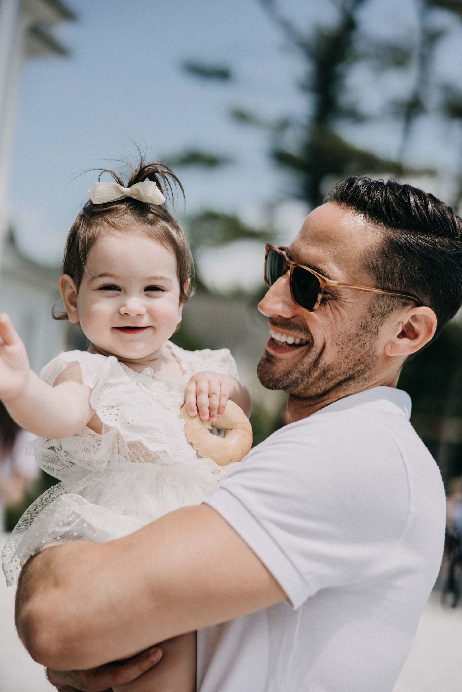 A candid outdoor portrait of a toddler in a white lace dress being held by someone wearing a white t-shirt and sunglasses.