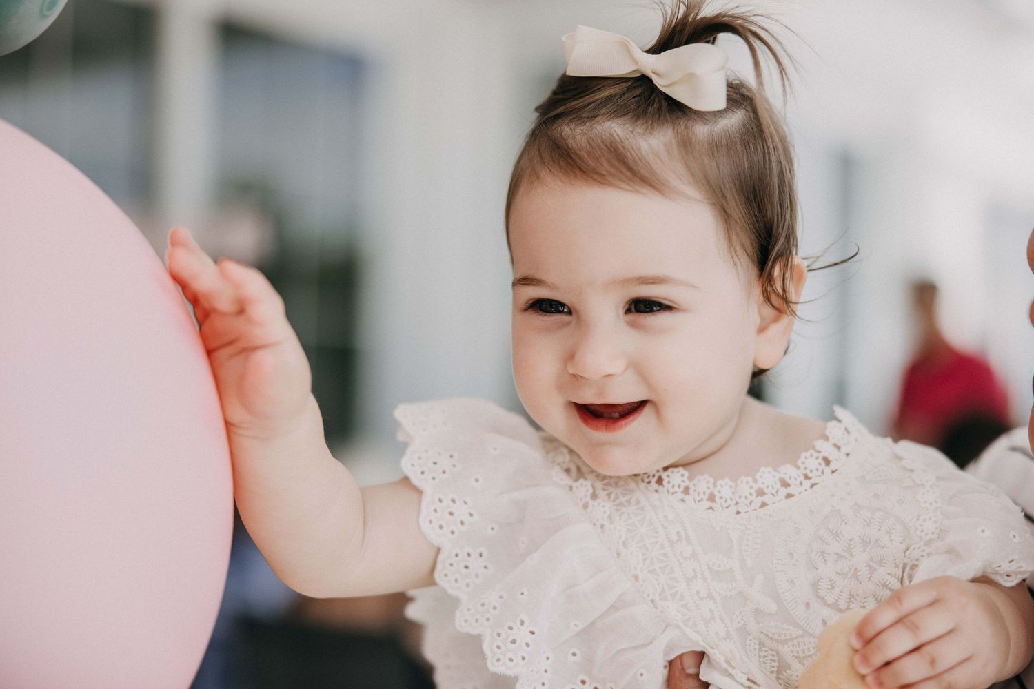 A joyful baby in white lace dress reaching for pink balloons at a party.