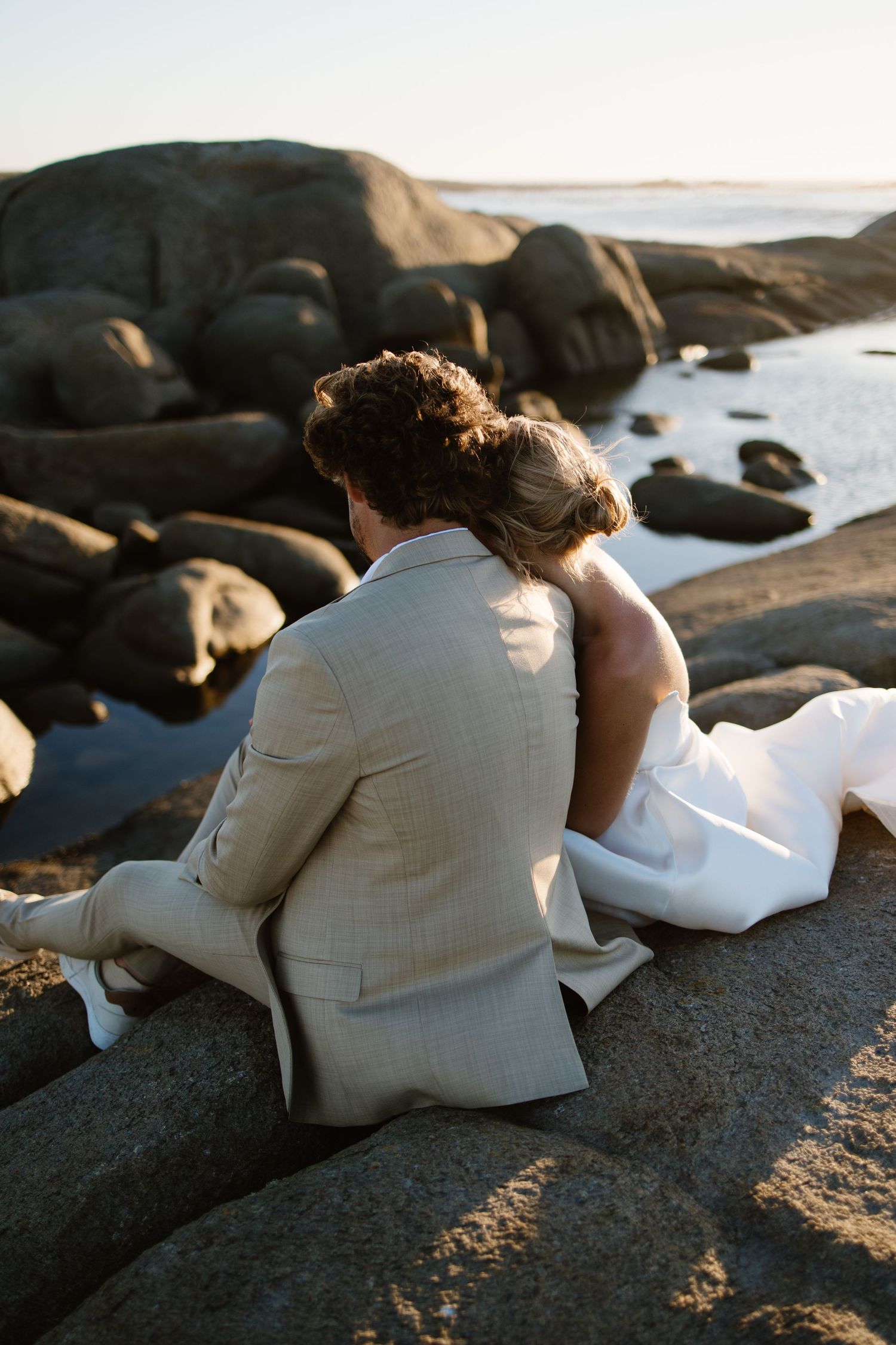 A bride and groom sit atop a boulder on the shore in Jacobs Bay, embracing as they watch the sun set over the ocean.