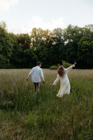 A couple runs playfully through a tall grass meadow at sunset.