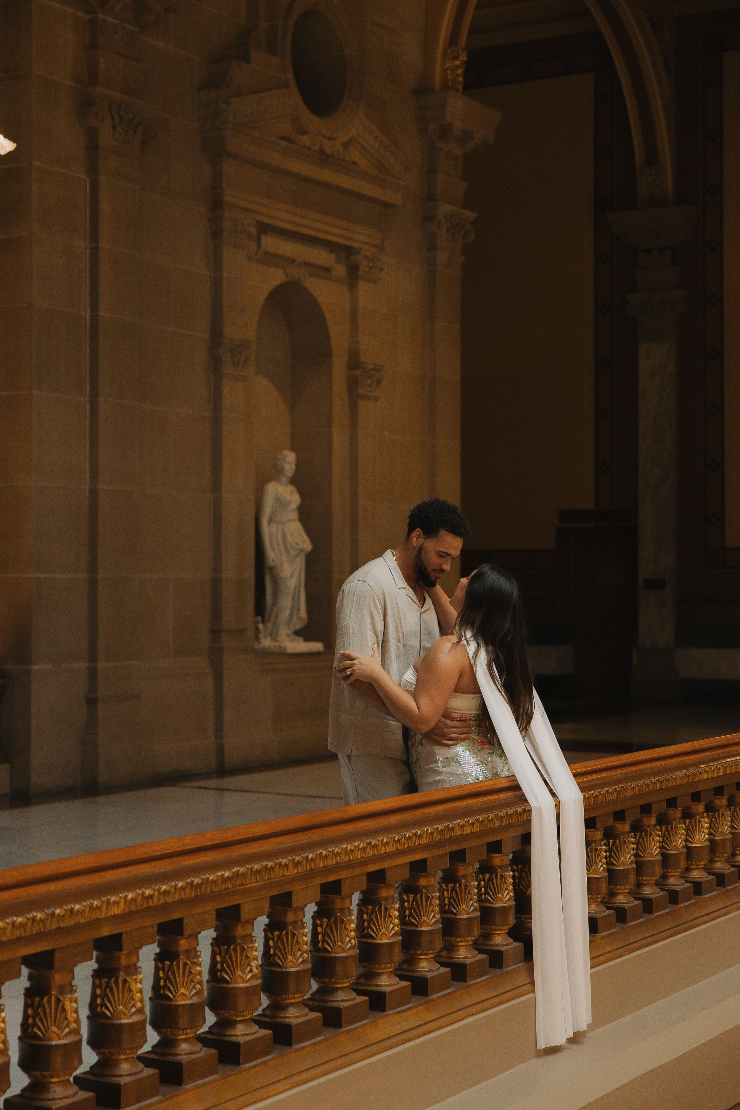 Couple sharing a romantic kiss on an ornate balcony inside a grand classical building with marble statue.