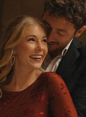 A smiling blonde woman in a red dress shares a romantic moment with a curly-haired man in a dark suit.