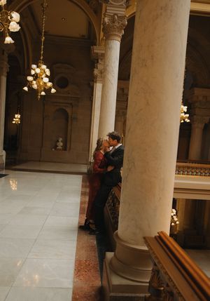 Couple sharing a romantic moment between marble columns in an elegant chandelier-lit hall.