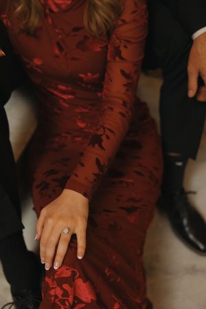 Woman in red floral dress displaying diamond engagement ring while seated beside a man in black suit.