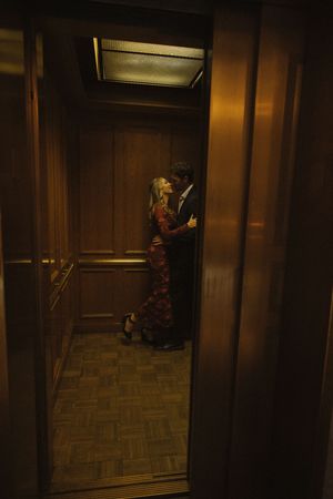Couple sharing a romantic kiss inside a warmly lit wooden elevator with parquet flooring.