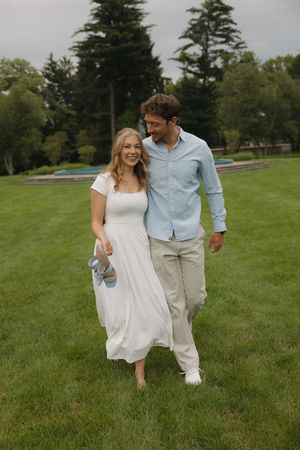A couple walks barefoot on lush green grass, woman in white dress holding shoes, man in blue shirt with arm around her.