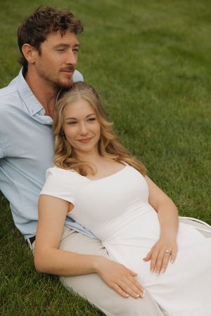 A pregnant woman in a white dress sits on grass with her partner, both smiling peacefully outdoors.