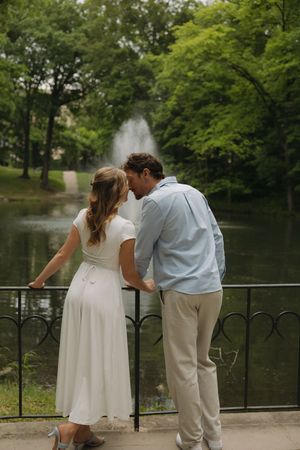 Couple standing by ornate railing in lush park, facing fountain on pond, woman in white dress, man in blue shirt.