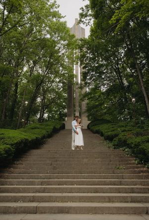 Couple embracing on grand stone steps surrounded by lush green trees and a tall monument.