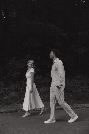 Black and white photo of a couple walking outdoors, woman in white dress smiling at man in light outfit near trees.