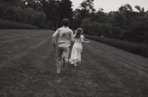 Black and white photo of a couple holding hands running across a lush green lawn surrounded by trees.