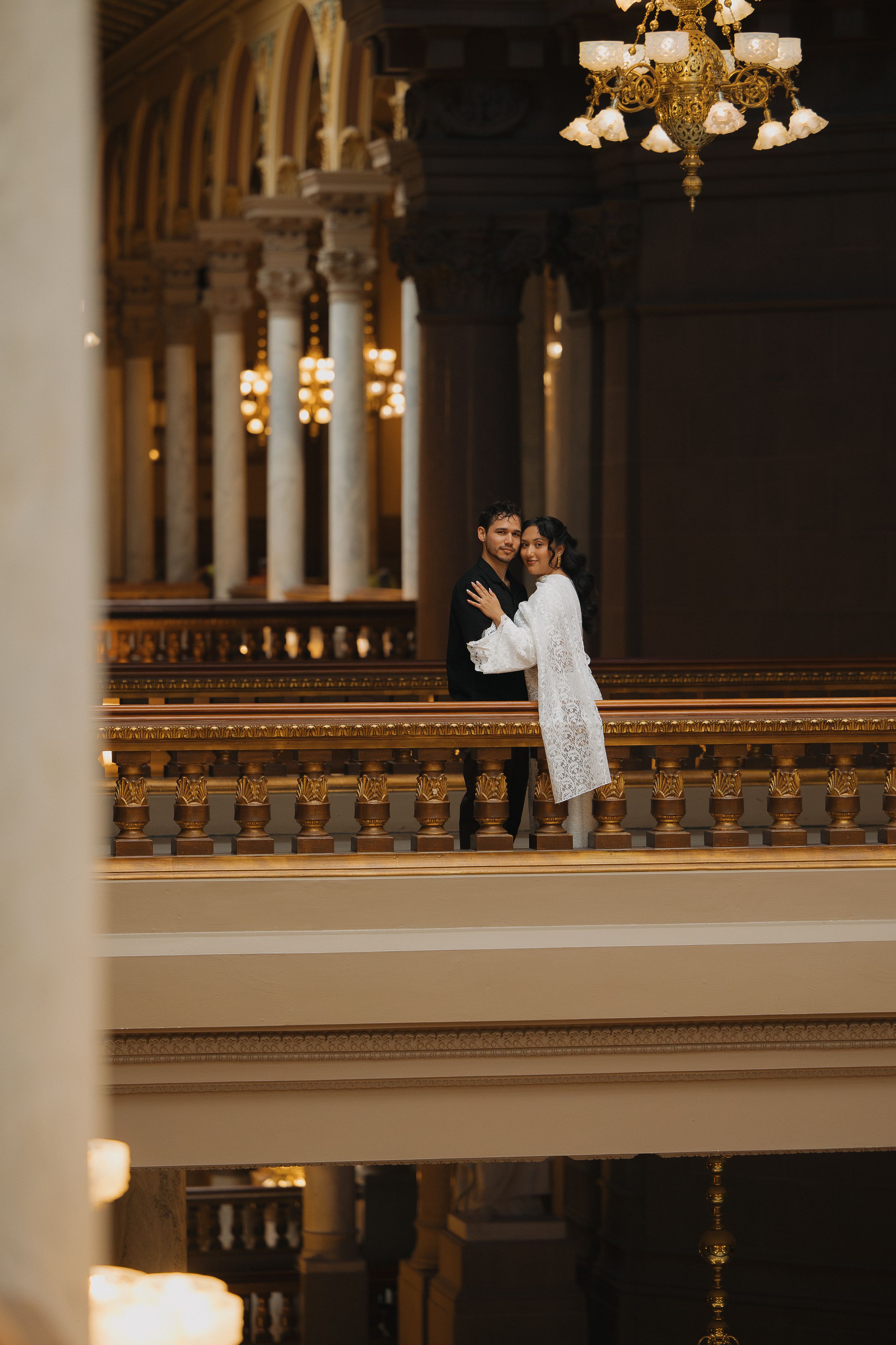 Couple embracing in elegant grand hall with ornate gold railings and chandeliers during wedding photography session.