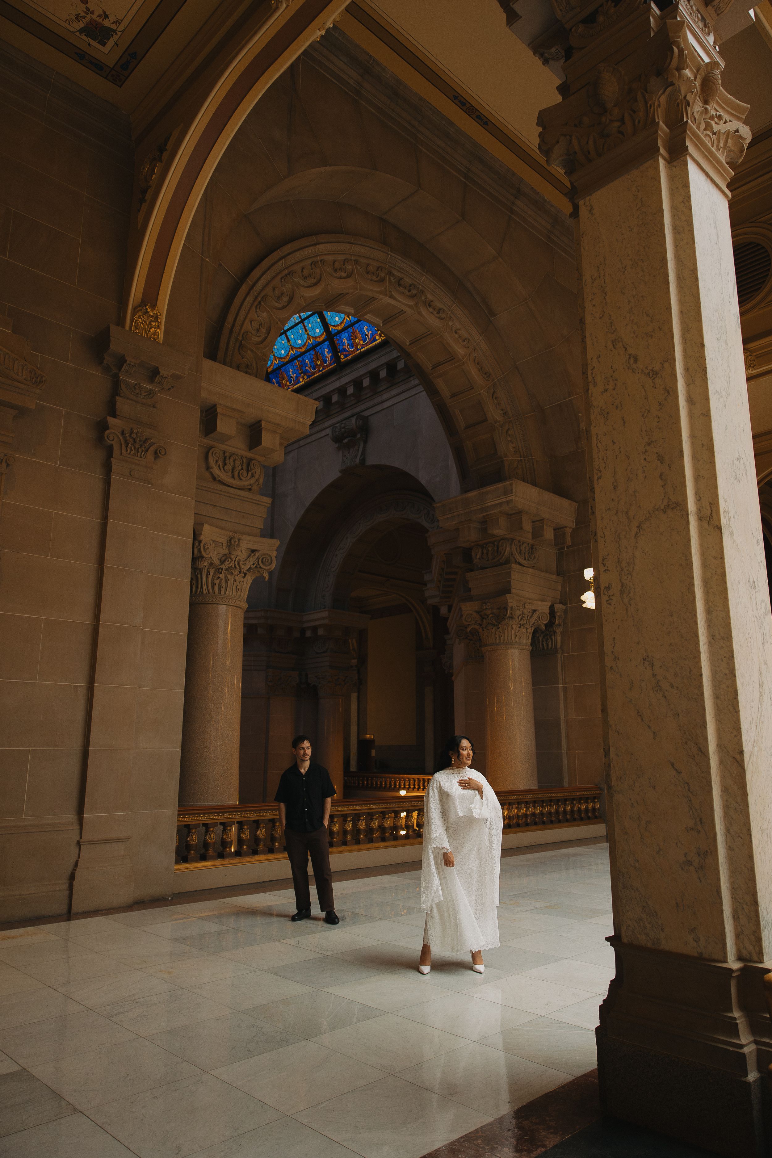 Couple in elegant wedding attire standing in a grand architectural hall with ornate arches and stained glass windows.