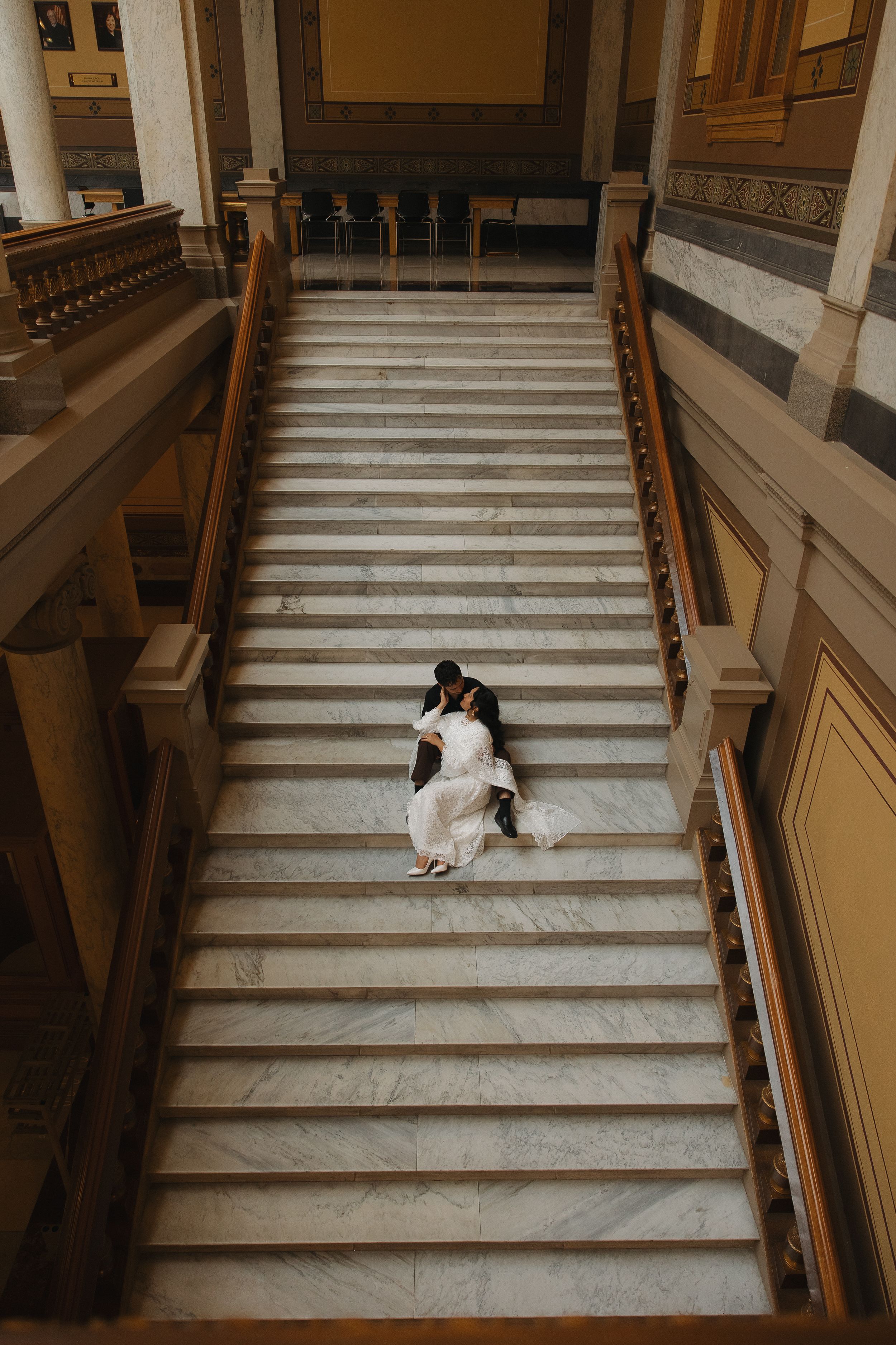 Bride and groom sitting together on grand marble staircase in elegant building, aerial view wedding portrait.