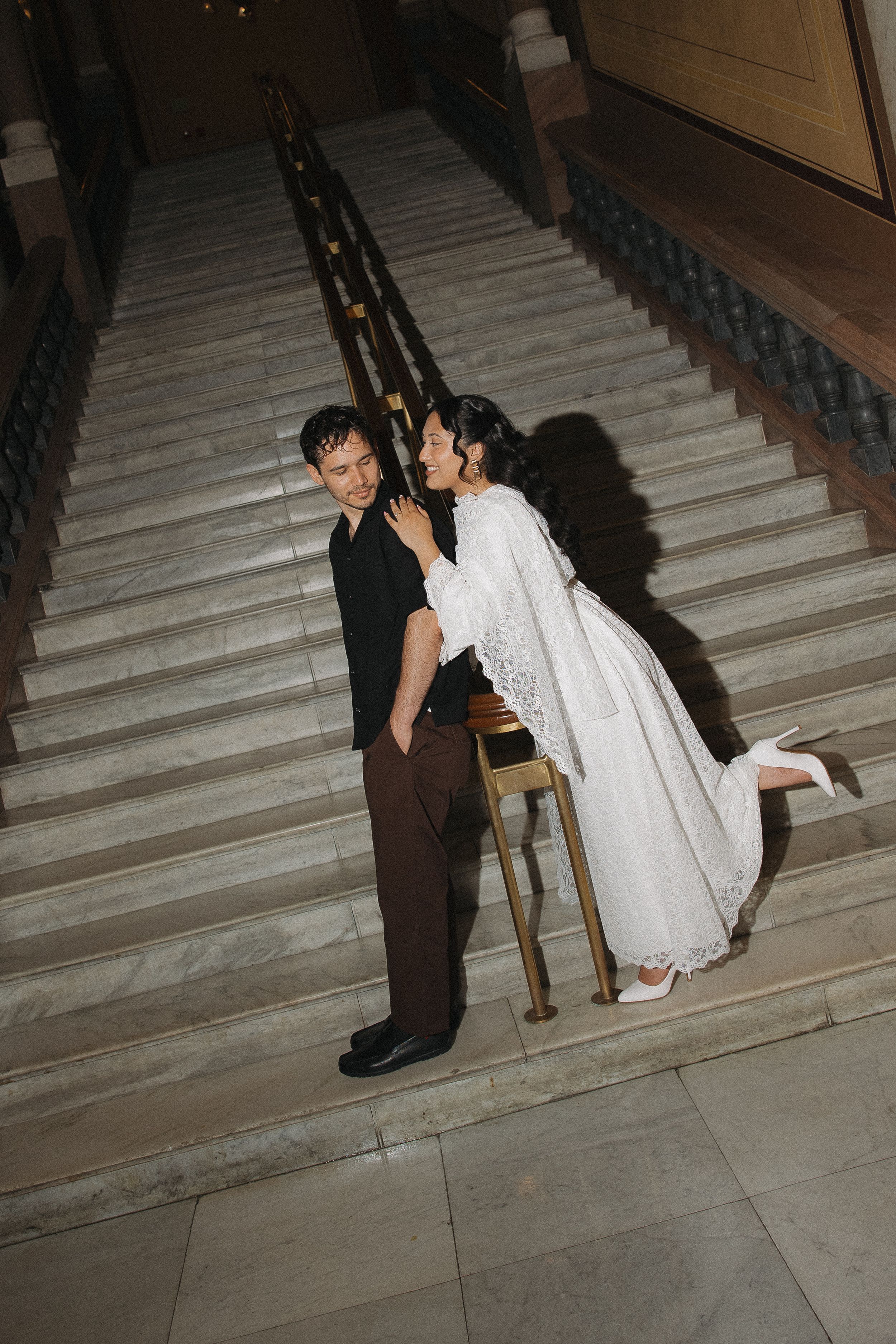 Bride in white lace gown playfully poses with groom on elegant marble staircase during wedding photo session.