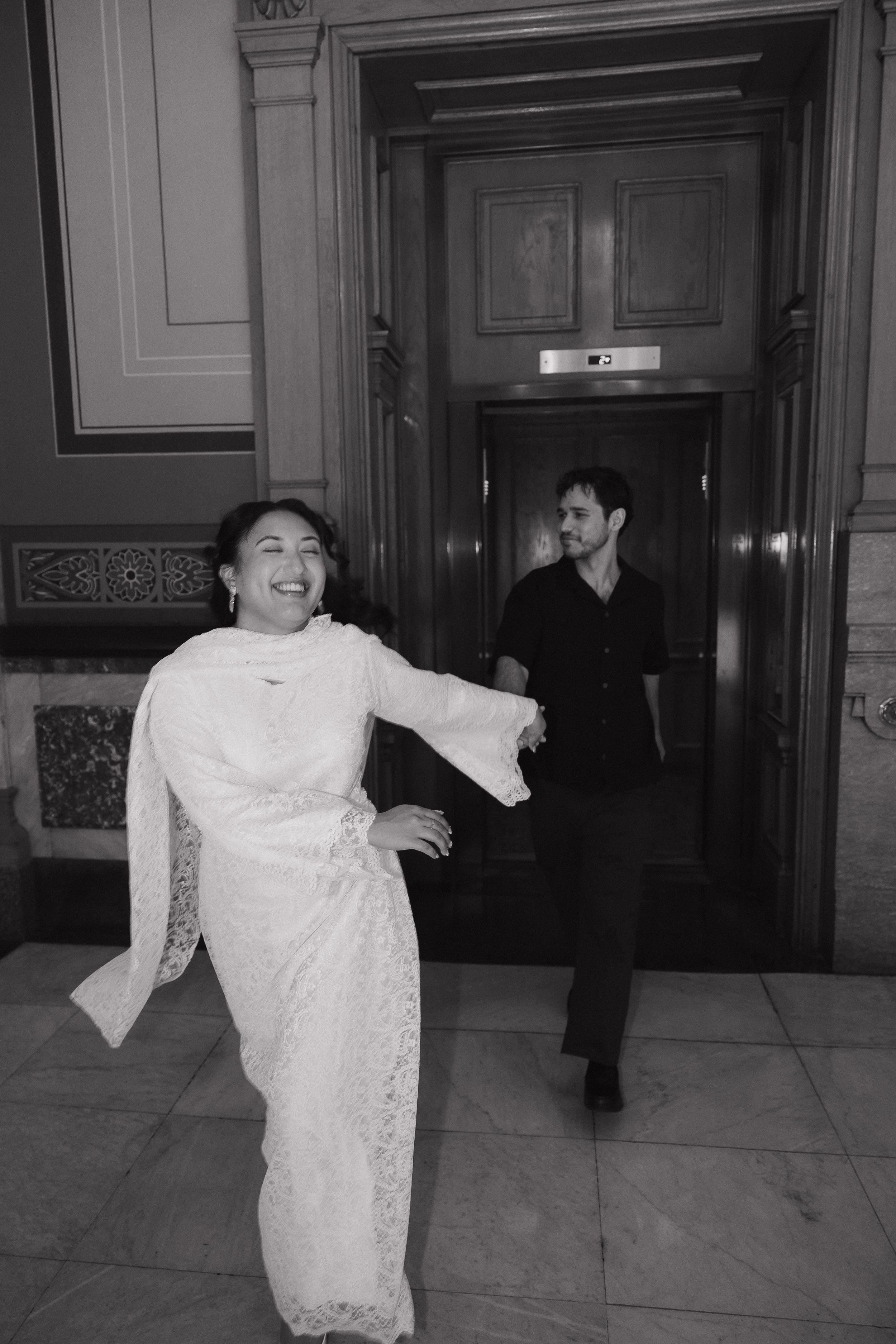 Black and white photo of a joyful woman in white lace dress and man in black exiting elevator.