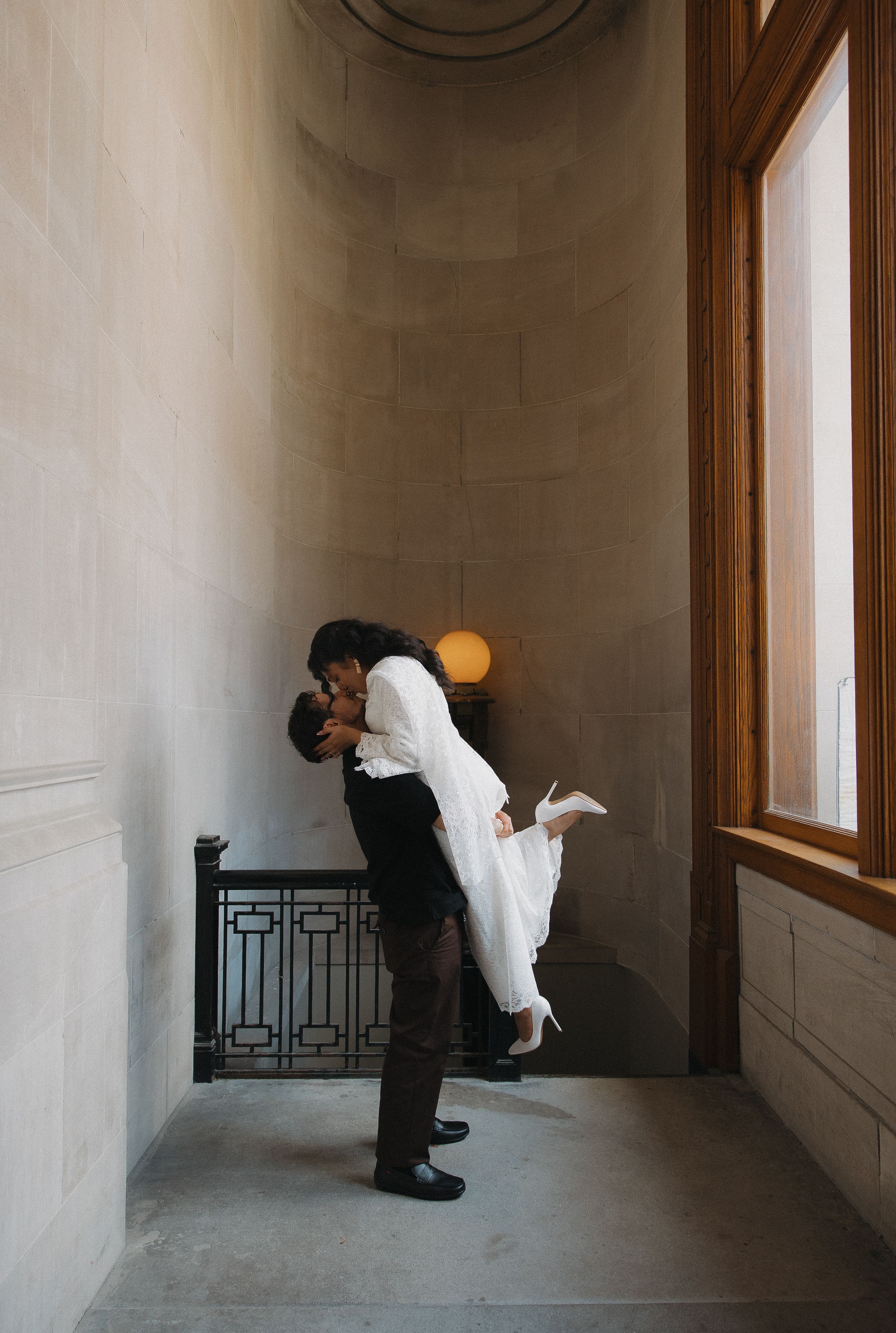 Groom lifts bride in white dress for a romantic kiss in an elegant stone hallway near a large window.