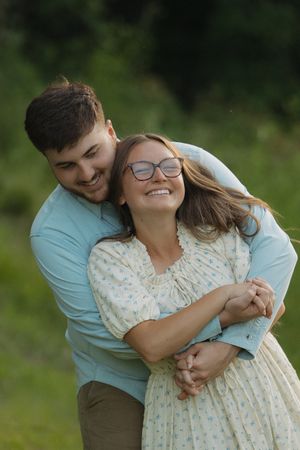 Happy couple embracing outdoors, woman in floral dress with glasses, man in blue shirt, smiling in green nature setting.