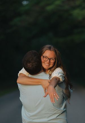 A smiling woman with glasses hugs a man outdoors during golden hour, with blurred green trees in the background.