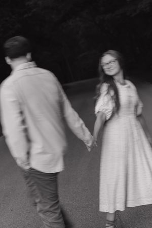 Black and white photo of a couple holding hands on a road, woman in dress smiling at camera.
