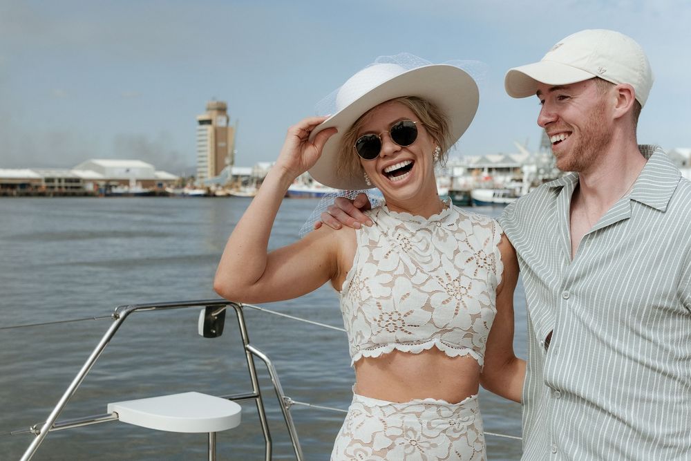 A laughing couple in summer outfits and hats enjoy a sunny boat ride with a waterfront skyline behind them.
