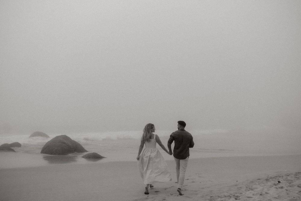 Two figures walk hand in hand along a misty beach with large rocks scattered in the foggy seascape.