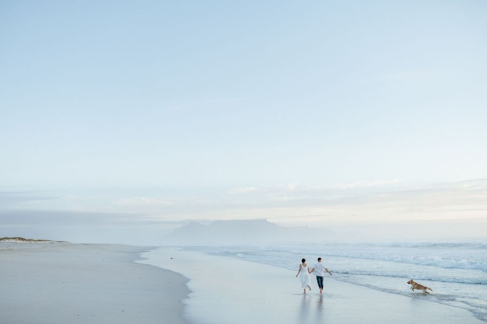 Couple holding hands walk along a wet sandy beach at dusk with a dog trotting nearby and soft pastel sky over the calm ocean.