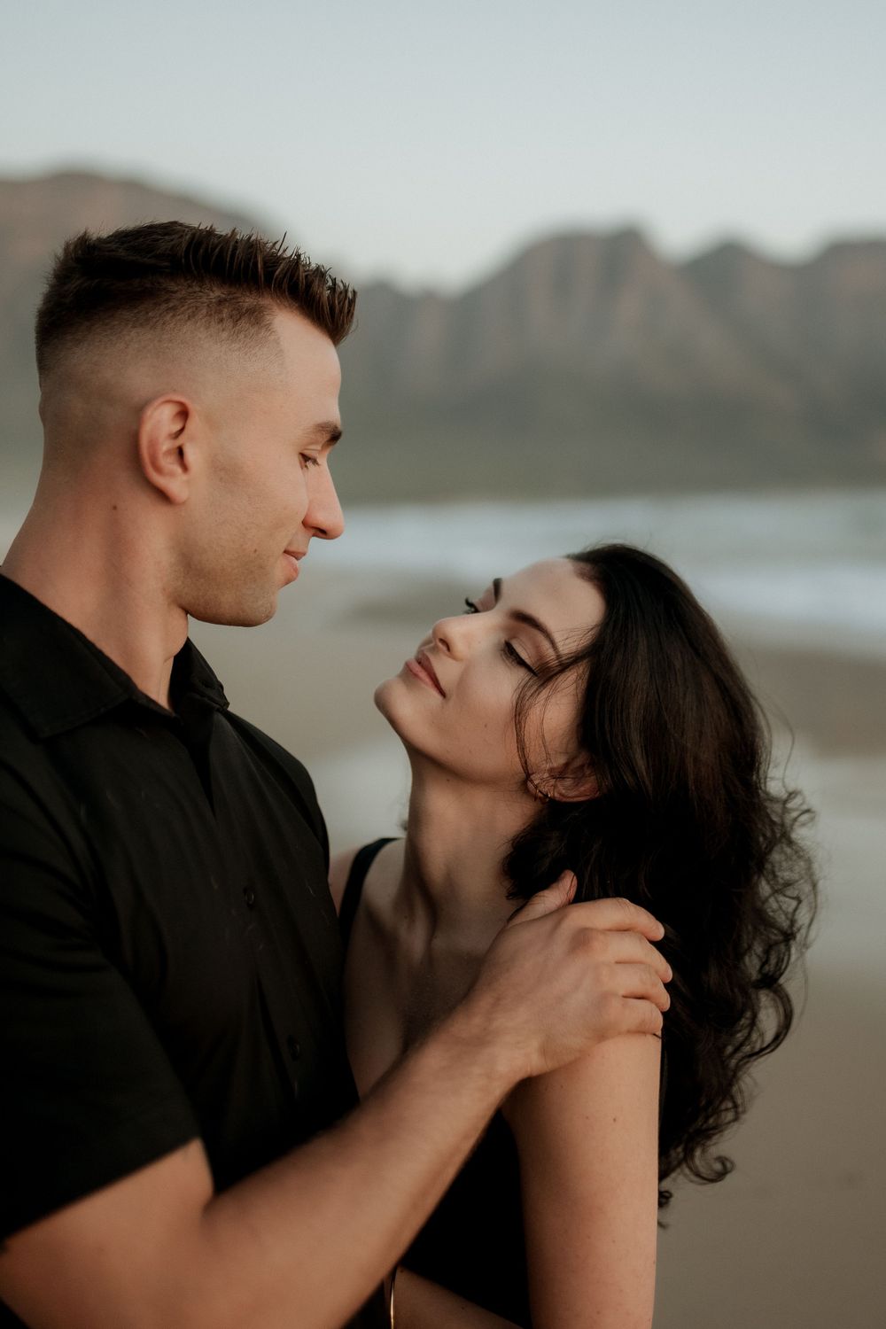 An intimate couple embraces on a beach at dusk with mountains visible in the background.