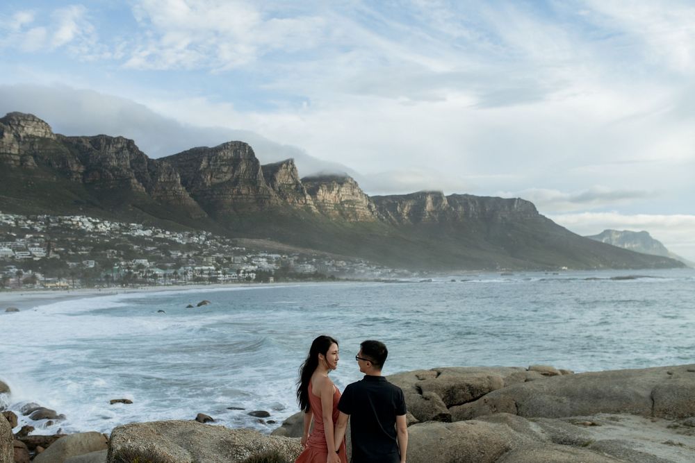Couple standing on rocky shore with ocean waves and mountain backdrop at Camps Bay, Cape Town.