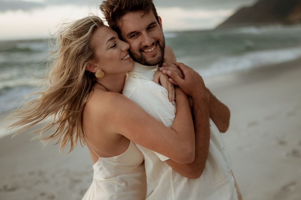 A loving couple embraces and laughs together on a windswept beach at sunset.