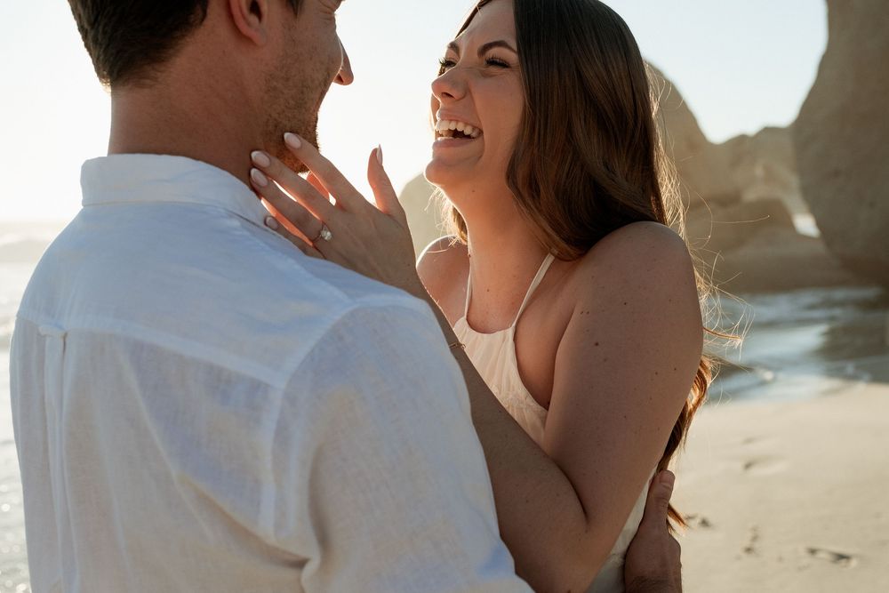Couple sharing a joyful moment on a beach at sunset, woman laughing and showing engagement ring.